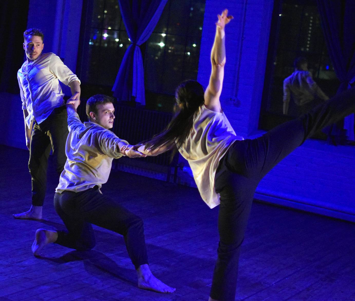 bathed in purple light three dancers  in white shirts and black pants cling to one another in a dramatic pose